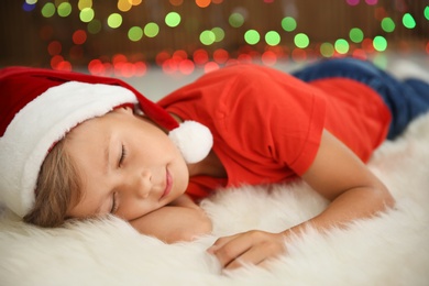 Photo of Cute little child in Santa hat sleeping on floor against blurred Christmas lights