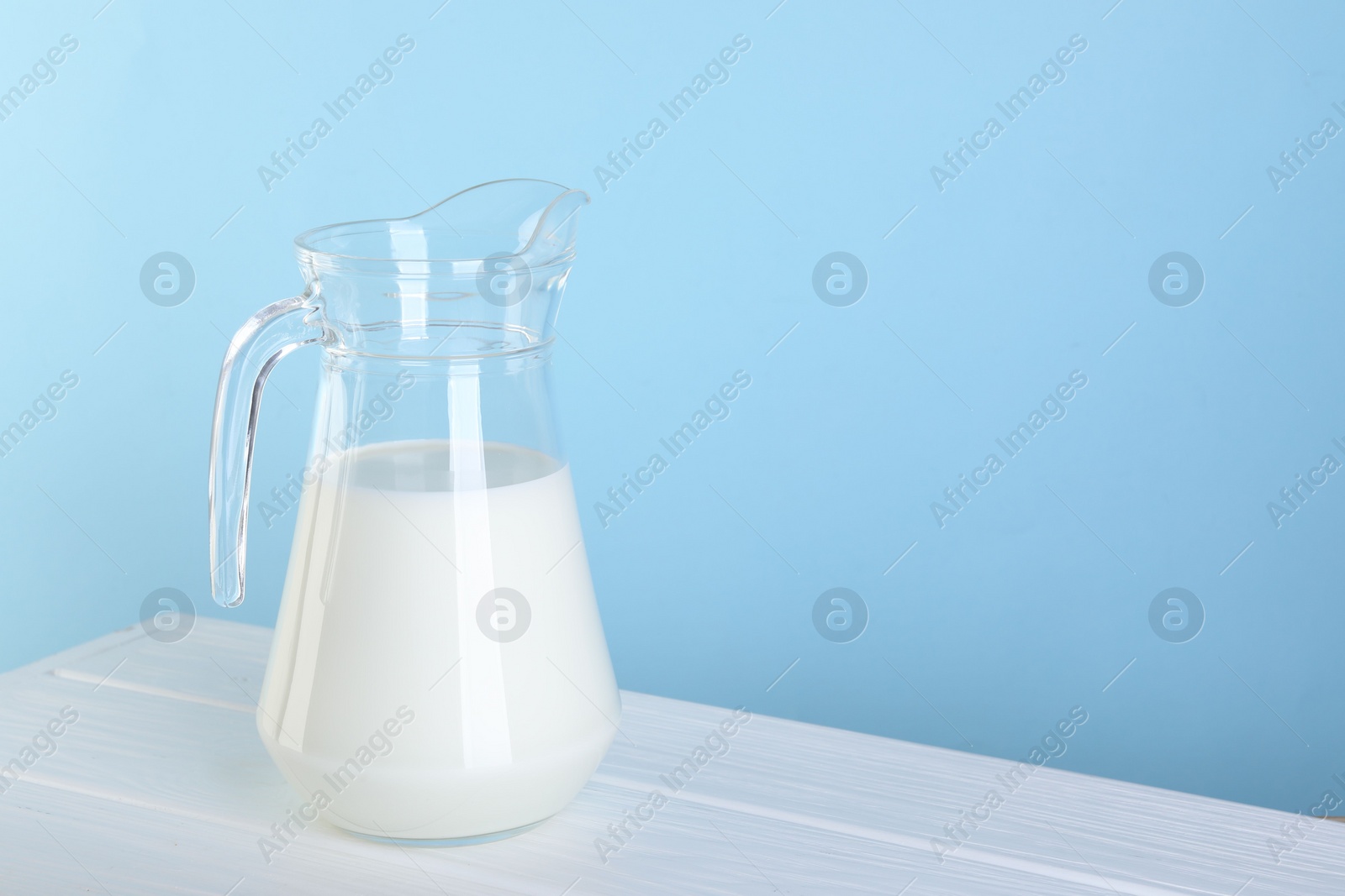 Photo of Jug of fresh milk on white wooden table against light blue background, space for text