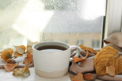 Cup of hot drink and autumn leaves near window on rainy day. Cozy atmosphere