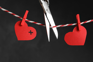 Photo of Red paper hearts on rope and scissors against black background. Composition symbolizing problems in relationship