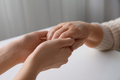 Photo of Young and elderly women holding hands together at white table indoors, closeup