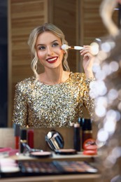 Beautiful makeup. Smiling woman applying powder with brush onto face in front of mirror indoors
