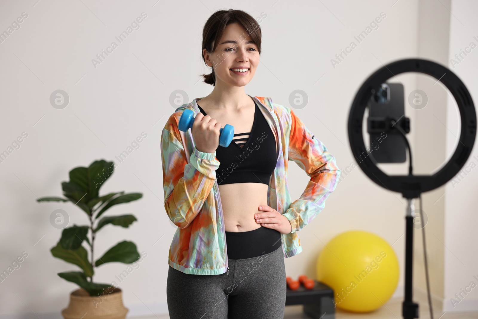 Photo of Happy sports blogger holding dumbbell while streaming online fitness lesson with smartphone at home