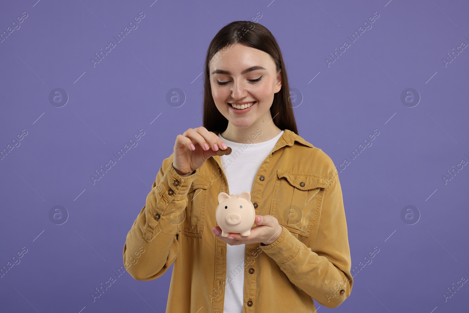 Photo of Happy woman putting coin into piggy bank on purple background