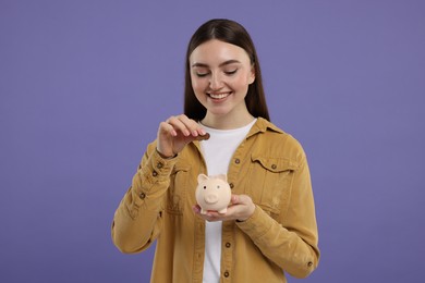 Happy woman putting coin into piggy bank on purple background