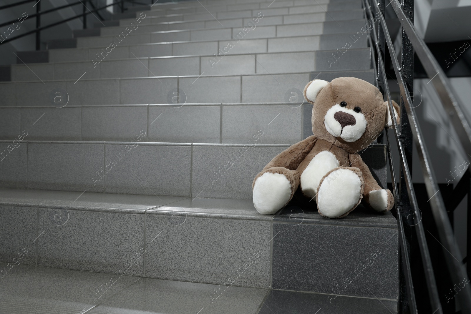 Photo of Lonely teddy bear on staircase near metal railing indoors, space for text