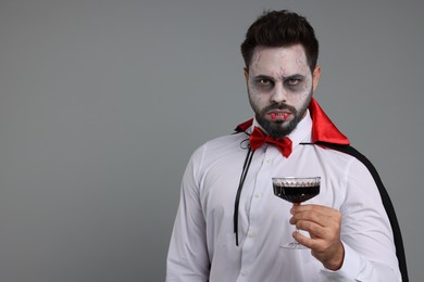 Photo of Man in scary vampire costume with fangs and glass of wine on light grey background, space for text. Halloween celebration
