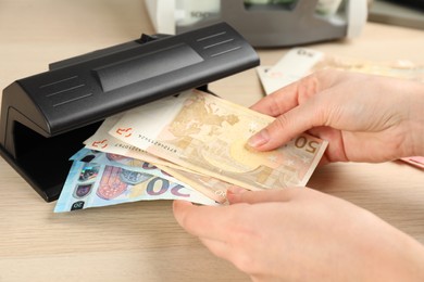 Woman checking Euro banknotes with currency detector at wooden table, closeup. Money examination device