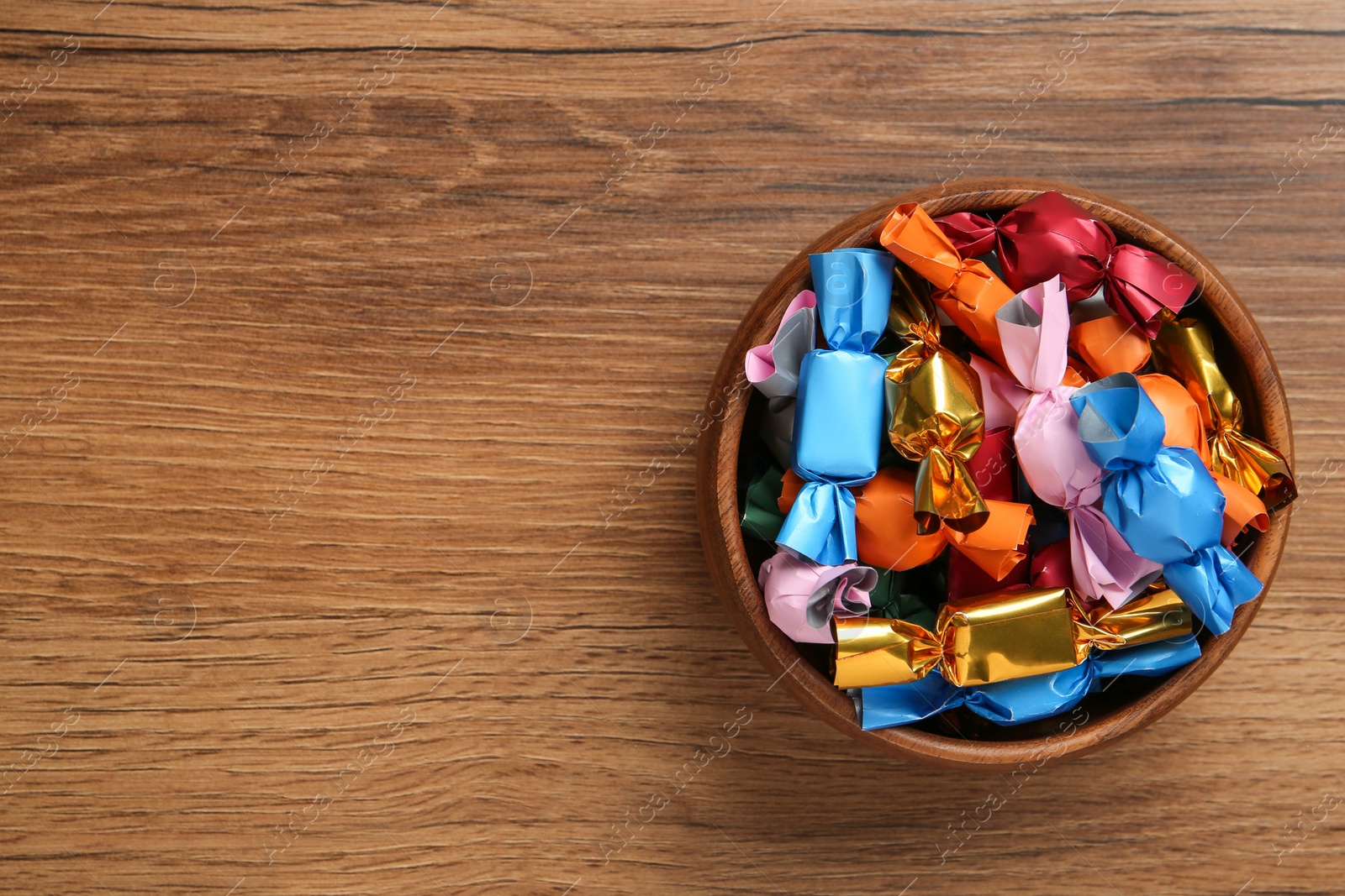 Photo of Bowl with candies in colorful wrappers on wooden table, top view. Space for text