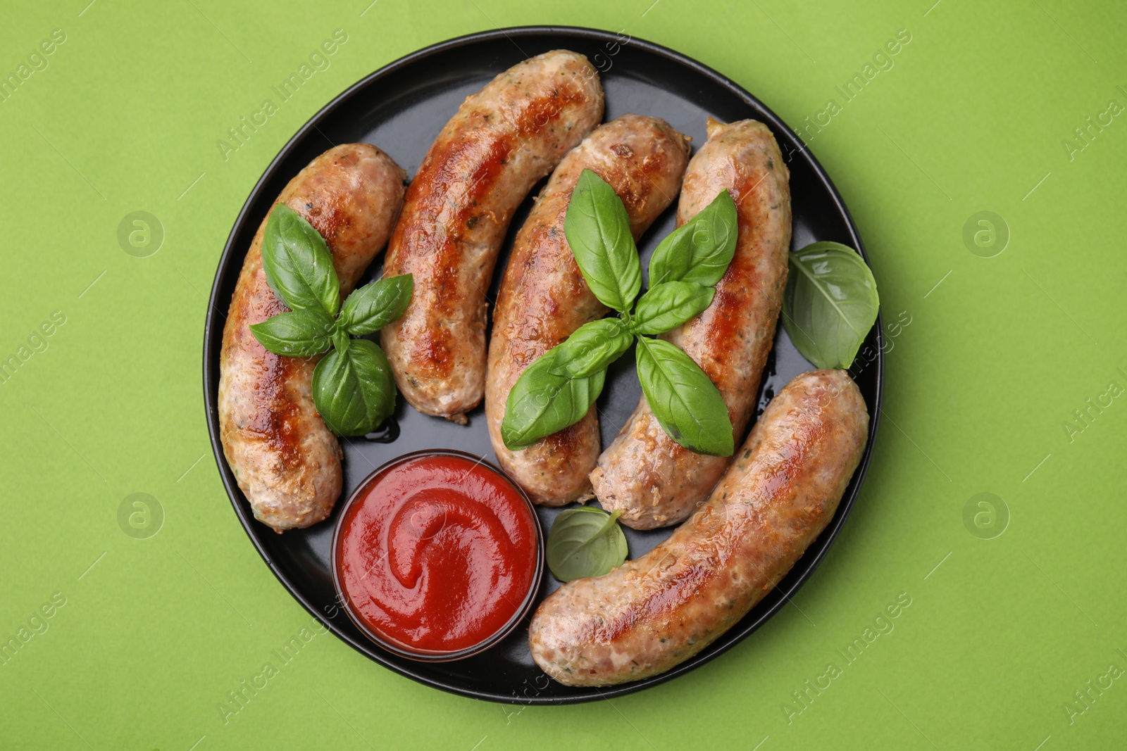 Photo of Plate with tasty homemade sausages, ketchup and basil leaves on green table, top view