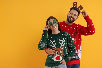 Photo of Happy young couple in Christmas sweaters, reindeer headband and party glasses on orange background. Space for text