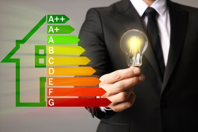 Image of Energy efficiency rating and man holding glowing light bulb on grey background, closeup