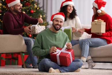 Photo of Christmas celebration in circle of friends. Happy young man opening gift at home, selective focus