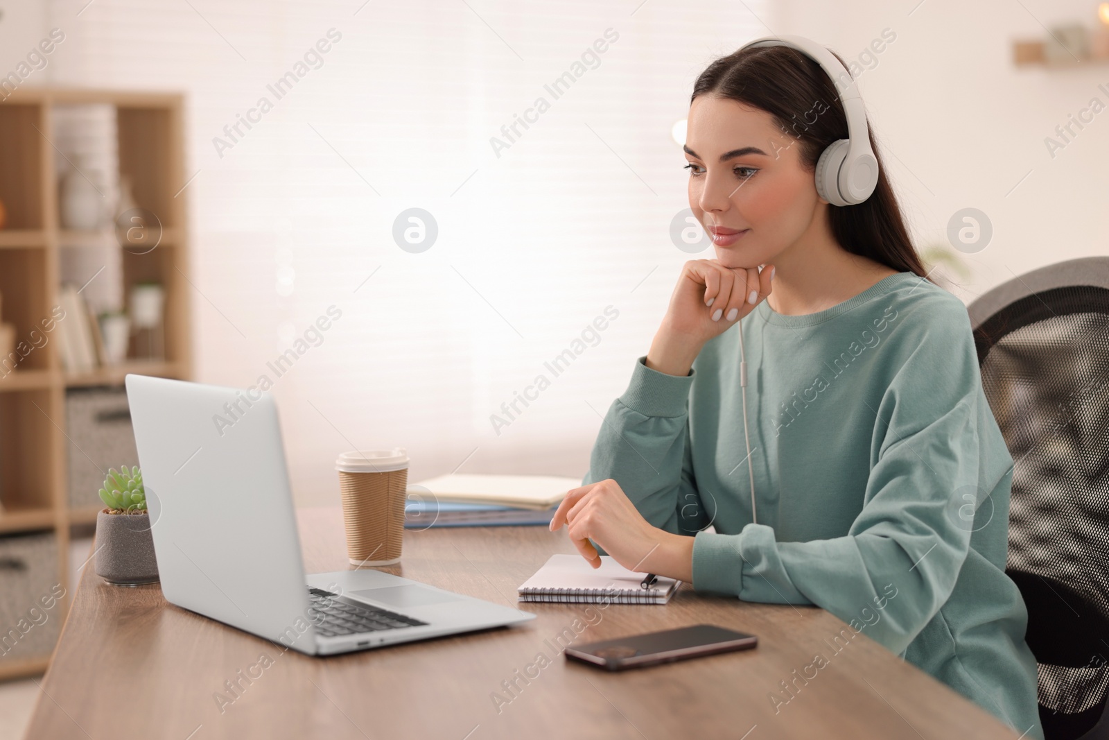 Photo of Young woman in headphones watching webinar at table in room