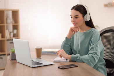 Photo of Young woman in headphones watching webinar at table in room