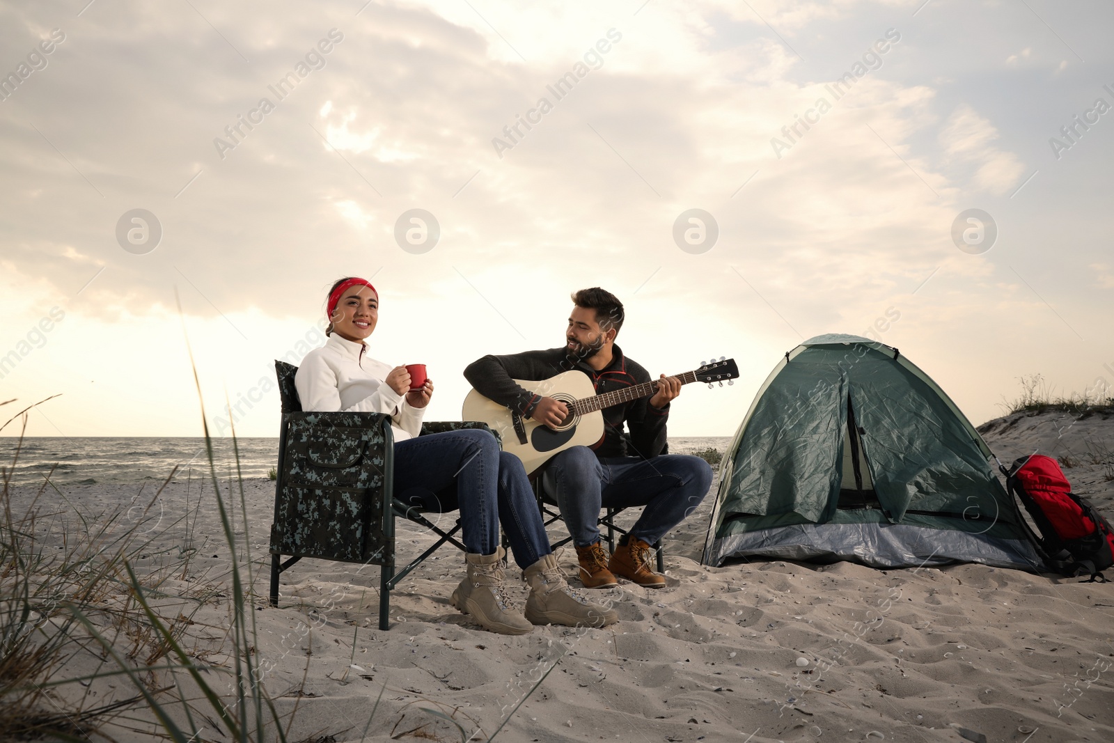 Photo of Young man playing guitar to his beloved girlfriend near camping tent on beach