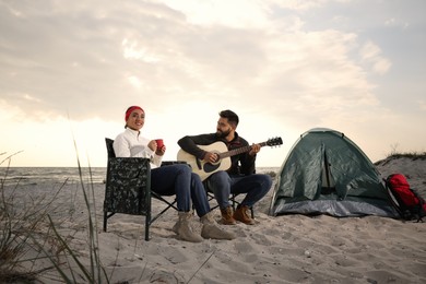 Young man playing guitar to his beloved girlfriend near camping tent on beach