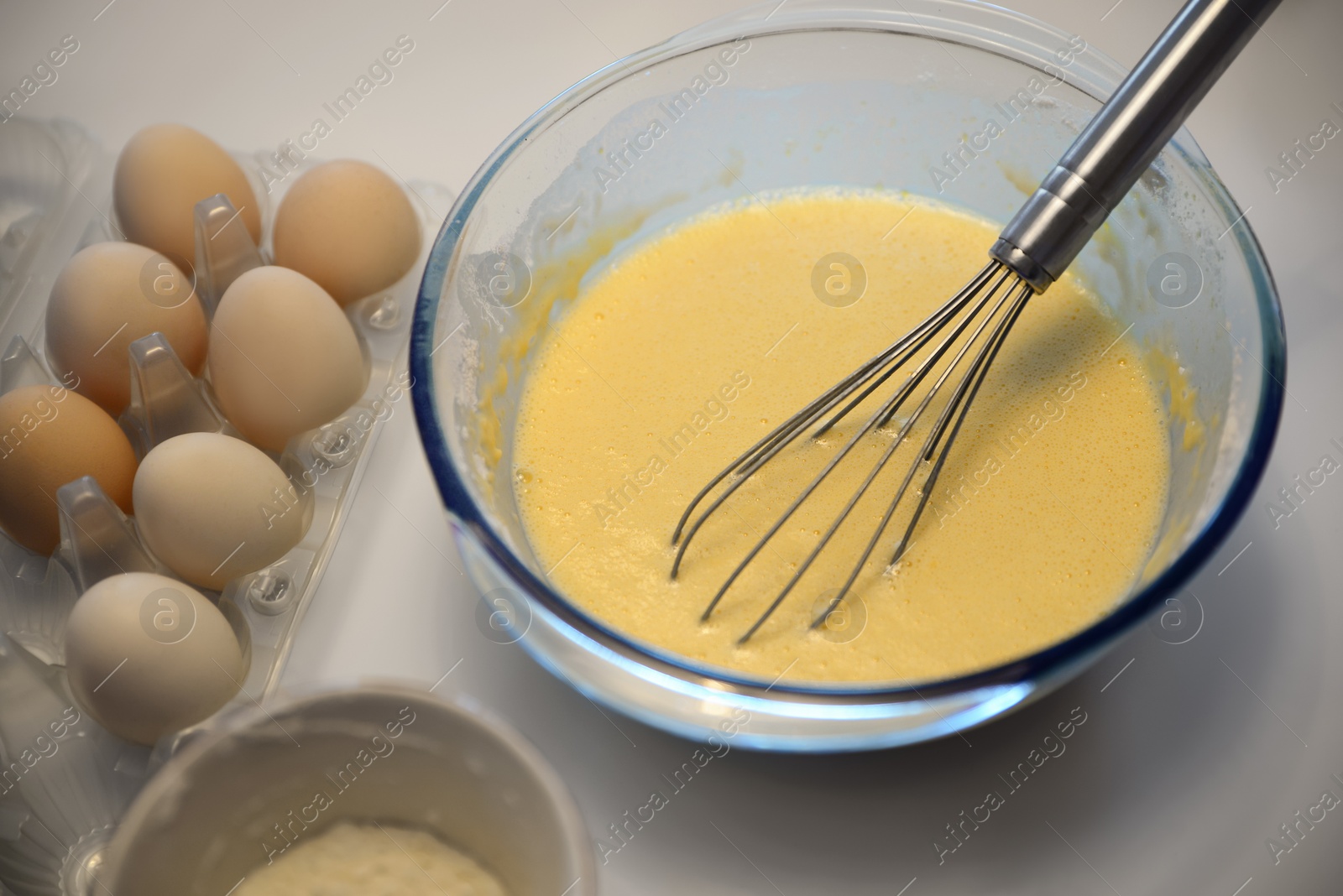 Photo of Glass bowl of crepe batter with whisk and ingredients on white table, closeup