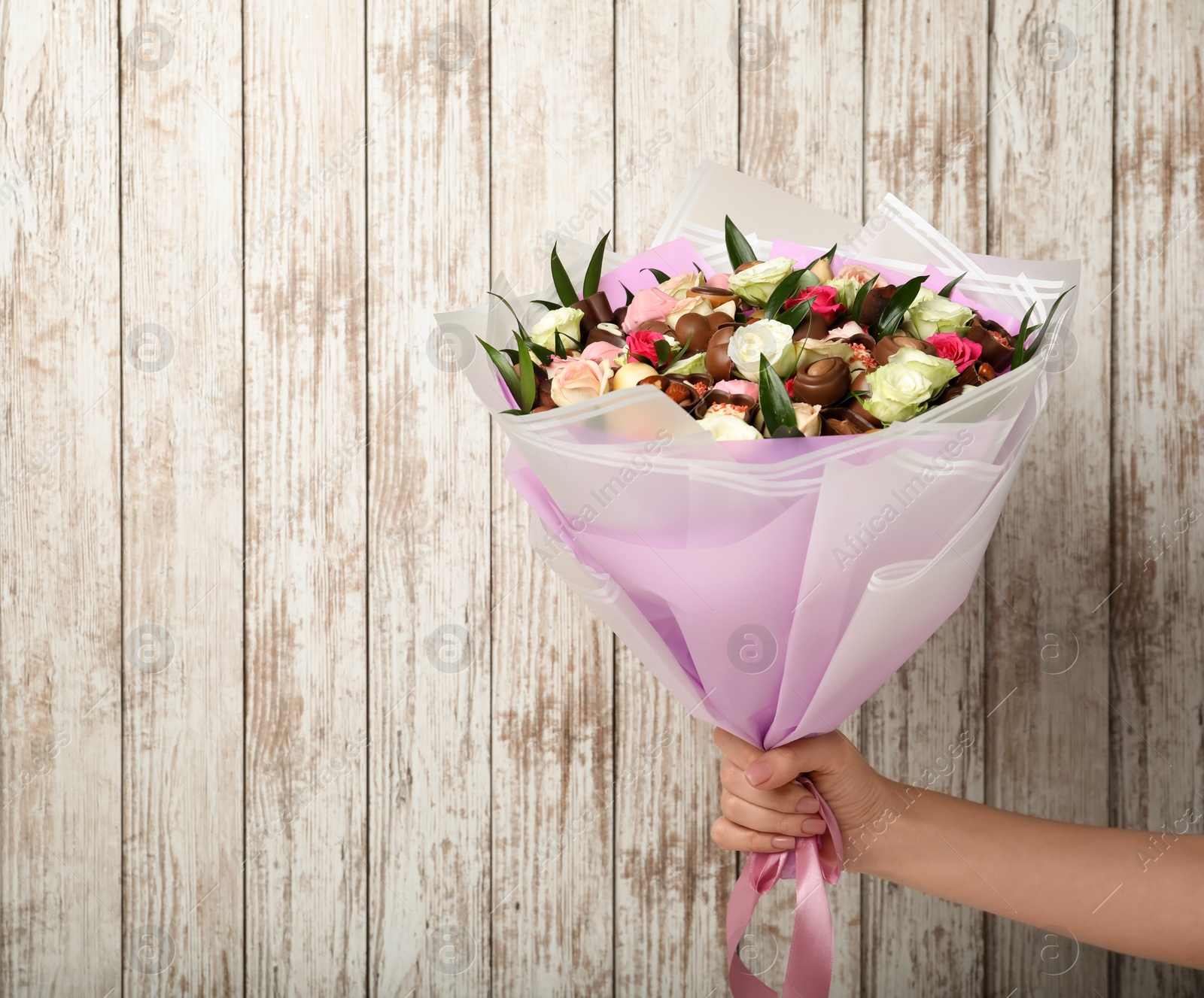 Photo of Woman holding beautiful bouquet of flowers and chocolate candies on white wooden background, closeup. Space for text