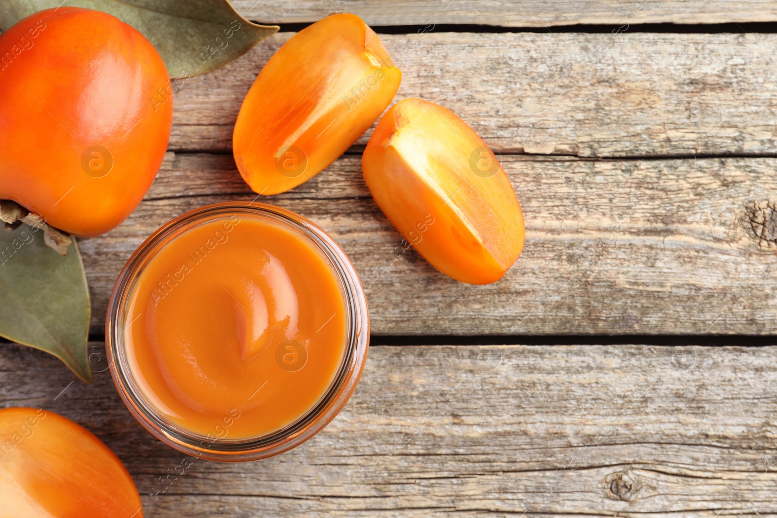 Photo of Delicious persimmon jam in glass jar and fresh fruits on wooden table, flat lay. Space for text