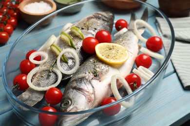 Photo of Glass baking tray with sea bass fish and ingredients on light blue wooden table, closeup