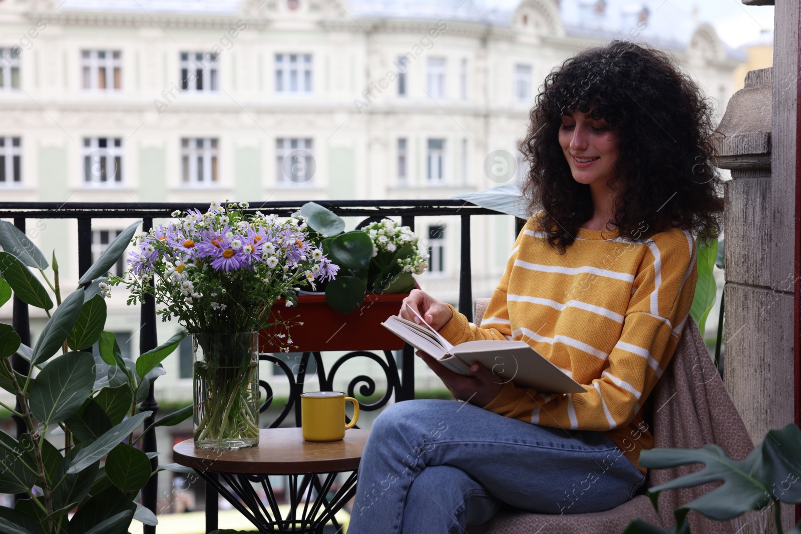 Photo of Young woman reading book at table on balcony with beautiful houseplants