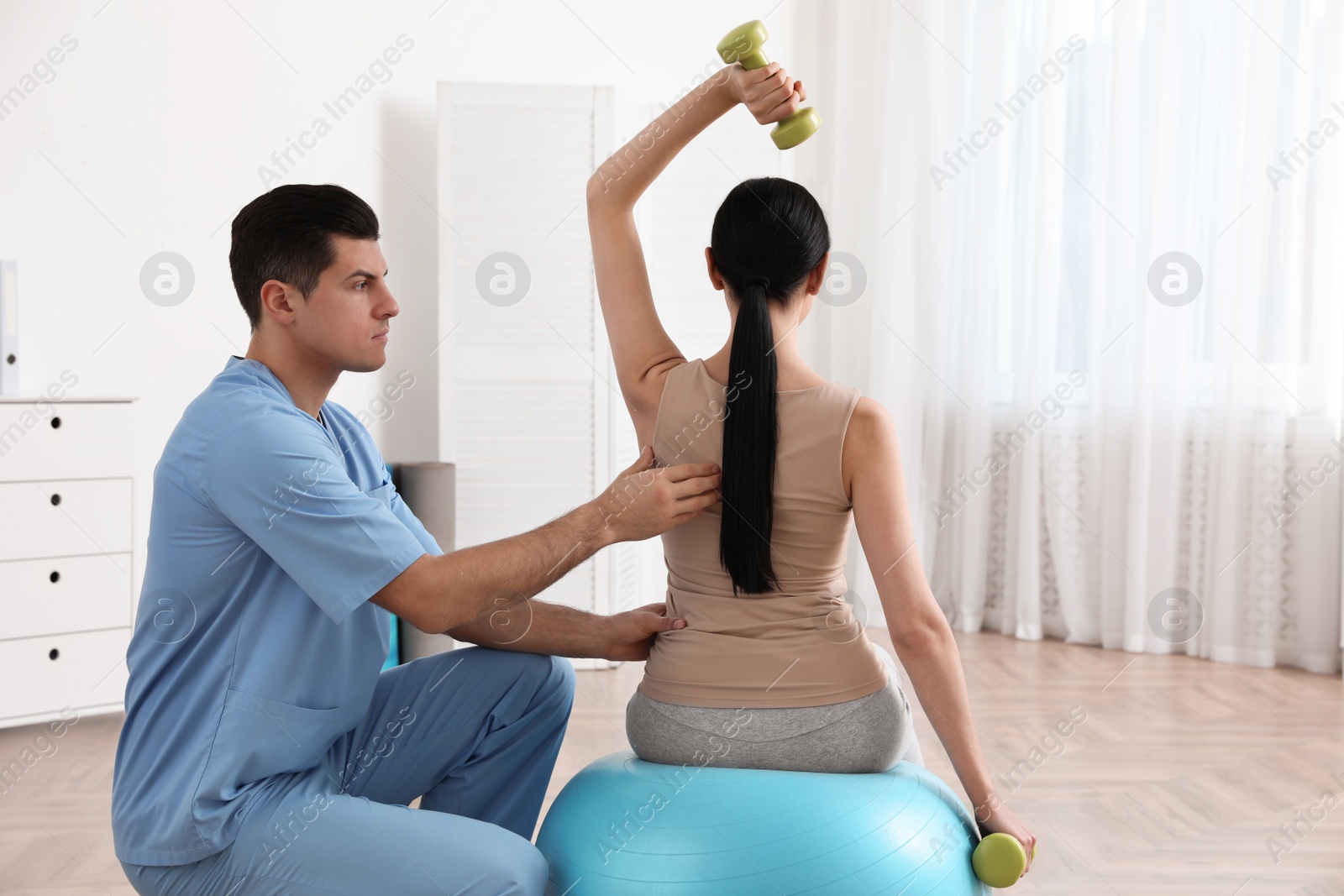 Photo of Orthopedist helping patient to do exercise with dumbbells in clinic. Scoliosis treatment