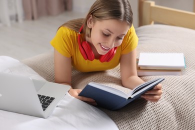 Photo of Online learning. Teenage girl reading book near laptop on bed at home