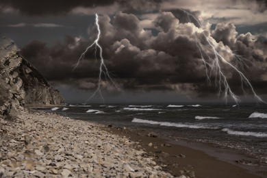 Image of Dark cloudy sky with lightnings over sandy beach with rocks and sea. Thunderstorm