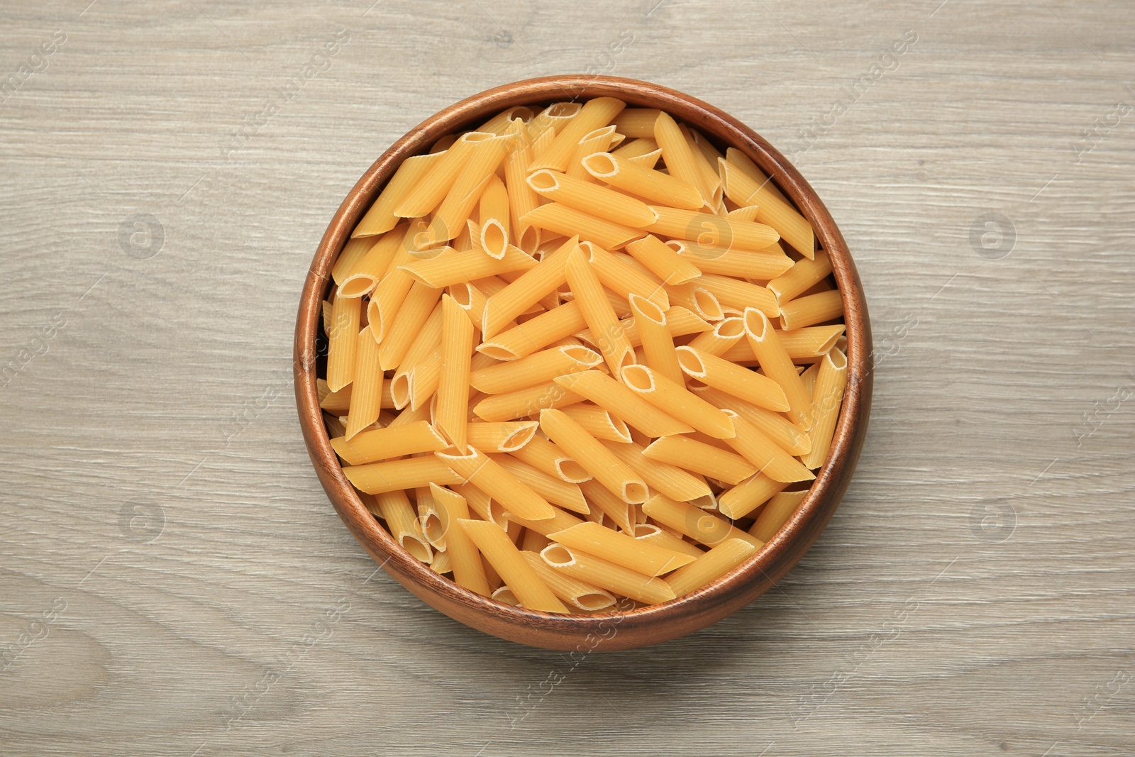 Photo of Raw penne pasta in bowl on light grey wooden table, top view