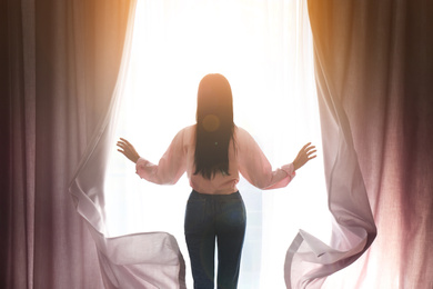 Image of Woman opening window curtains at home, back view