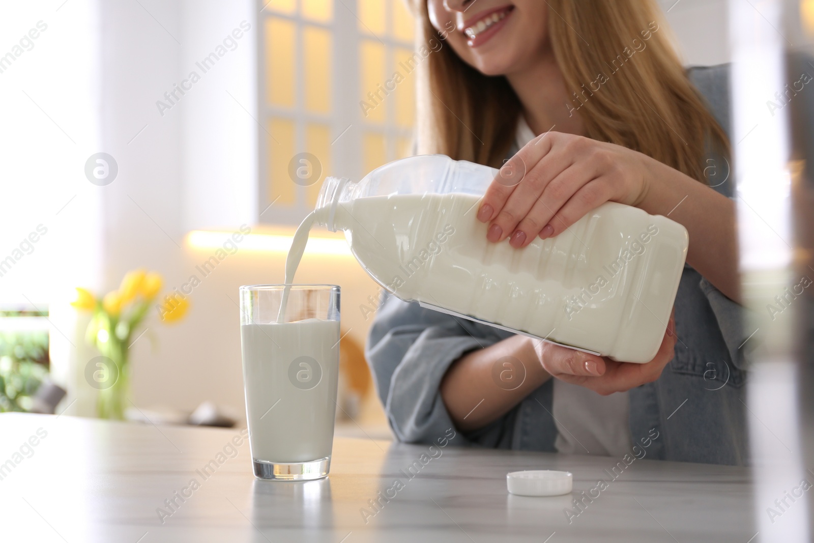 Photo of Young woman pouring milk from gallon bottle into glass at white marble table in kitchen, closeup