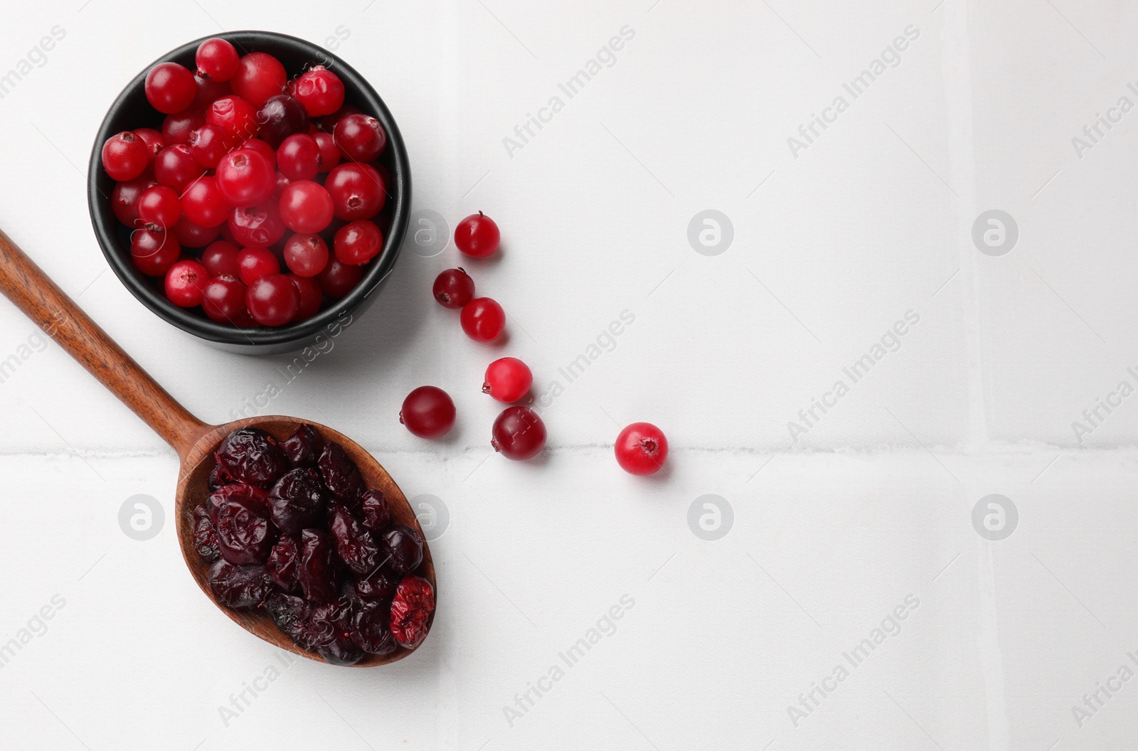 Photo of Tasty dried cranberries in spoon and fresh ones on white tiled table, top view. Space for text