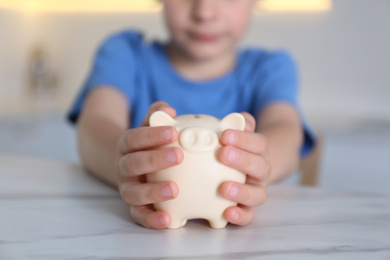 Little boy with piggy bank at marble table indoors, closeup