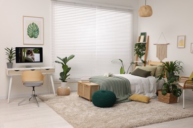 Comfortable bed, desk with computer and potted houseplants in stylish bedroom. Interior design