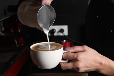 Photo of Barista pouring steamed milk from pitcher into cup of aromatic coffee in cafe, closeup