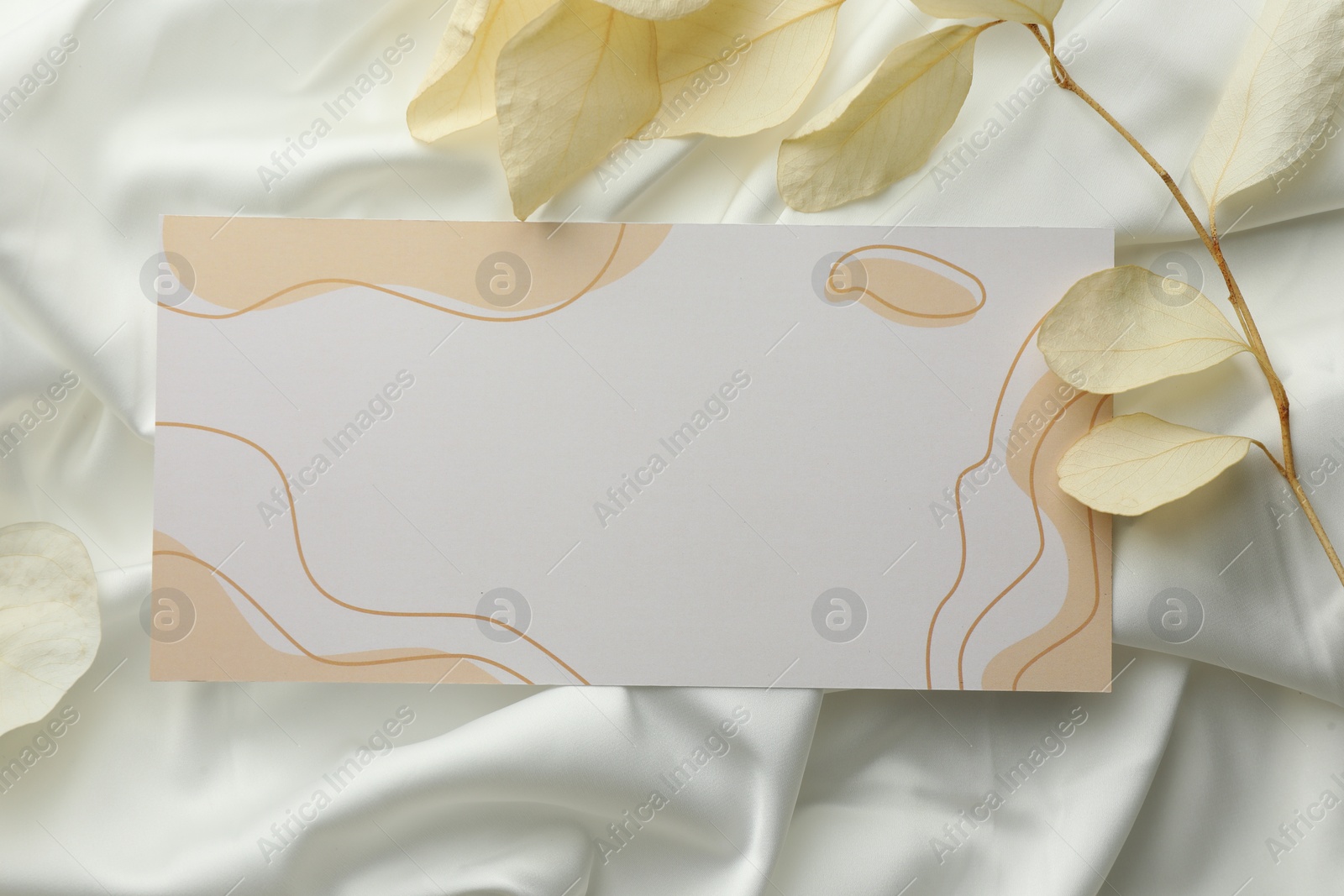 Photo of Blank invitation card and dry leaves on white fabric, flat lay