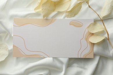 Photo of Blank invitation card and dry leaves on white fabric, flat lay