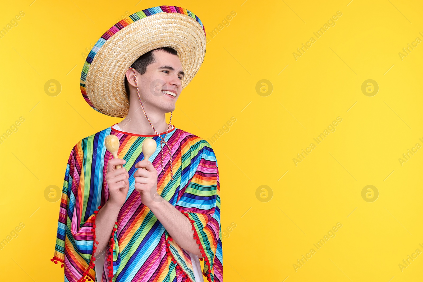 Photo of Young man in Mexican sombrero hat and poncho with maracas on yellow background. Space for text