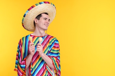 Young man in Mexican sombrero hat and poncho with maracas on yellow background. Space for text