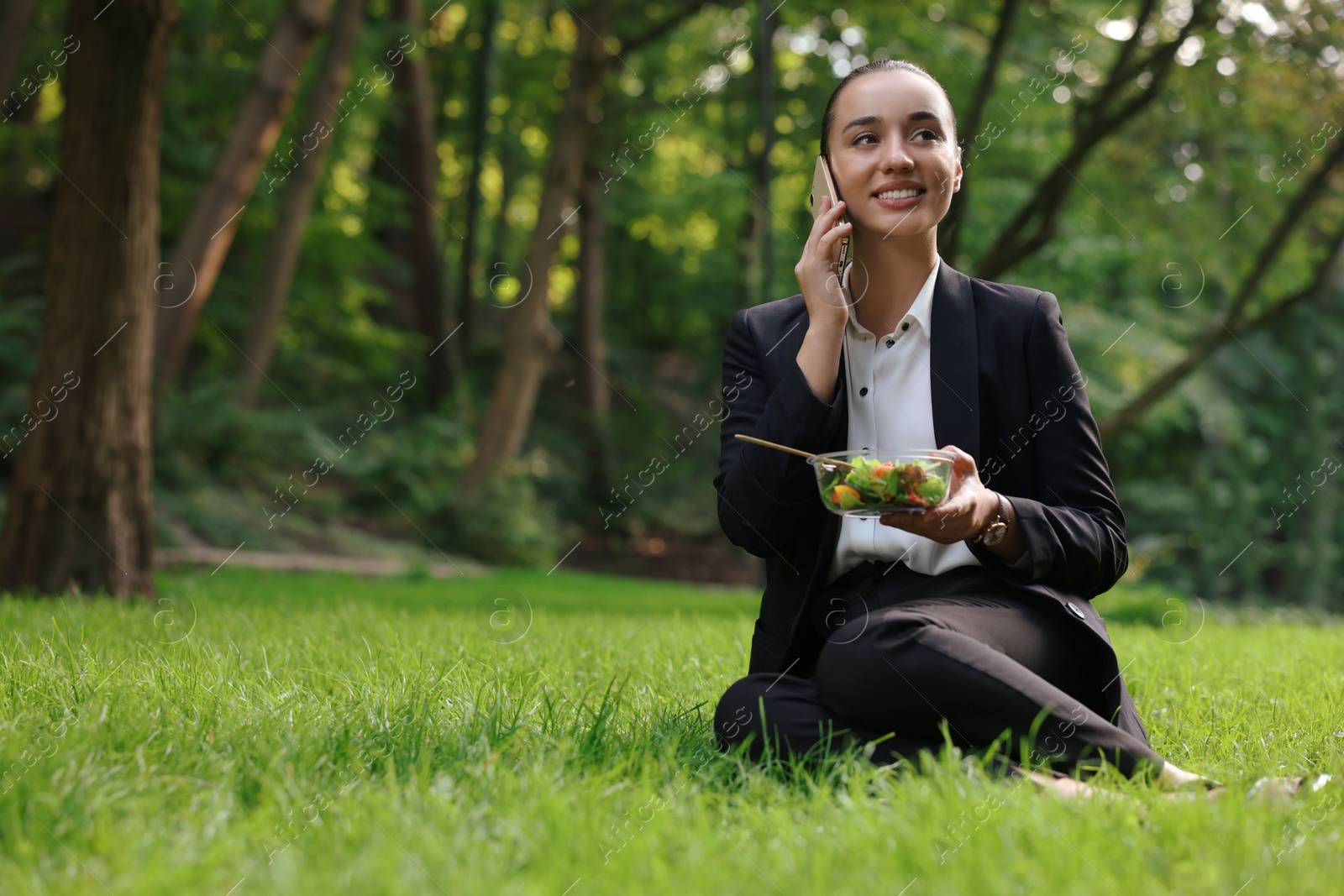 Photo of Lunch time. Happy businesswoman with container of salad talking on smartphone on green grass in park, space for text