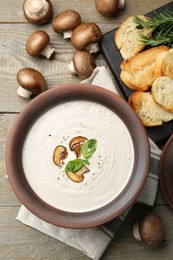 Photo of Tasty homemade mushroom soup in ceramic pot, croutons, fresh champignons and rosemary on wooden table, flat lay
