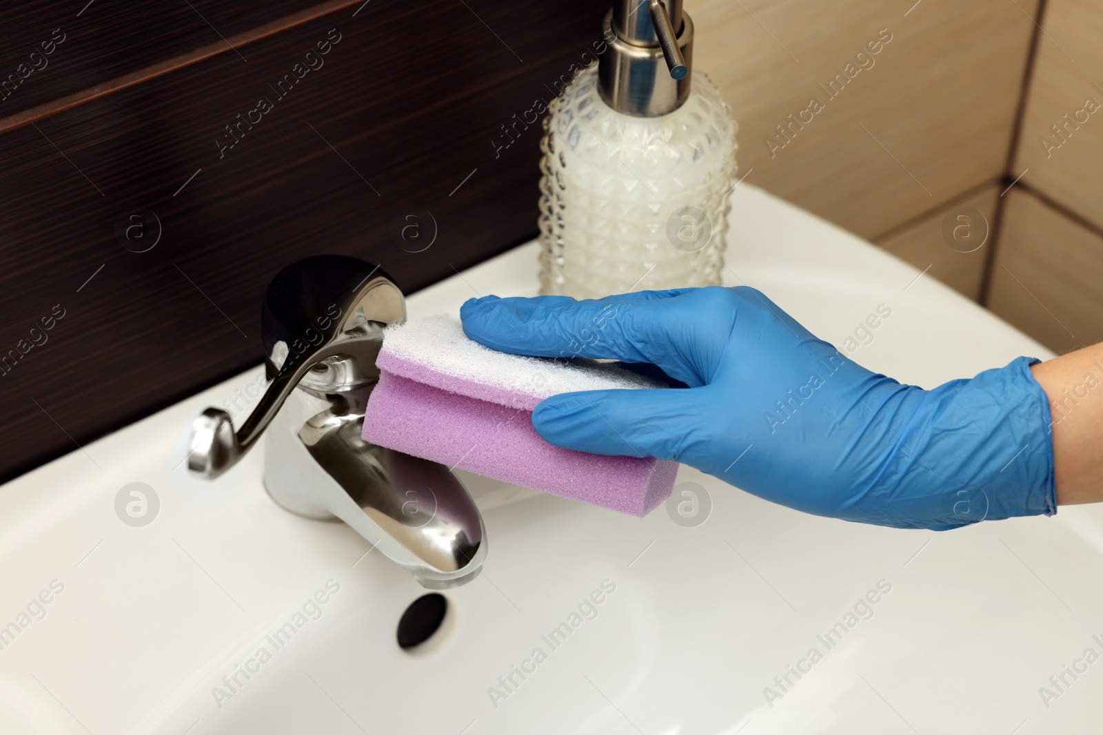 Photo of Woman in gloves cleaning faucet of bathroom sink with sponge, closeup