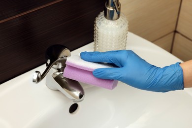 Photo of Woman in gloves cleaning faucet of bathroom sink with sponge, closeup