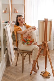 Beautiful young woman with drawing tools near easel at home