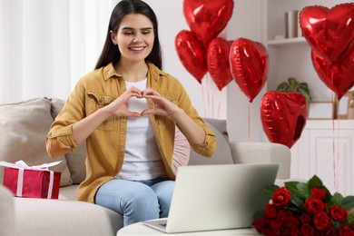 Photo of Valentine's day celebration in long distance relationship. Beautiful young woman making heart with hands while having video chat with her boyfriend via laptop