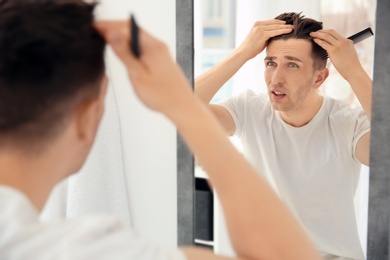 Young man with hair loss problem looking in mirror indoors