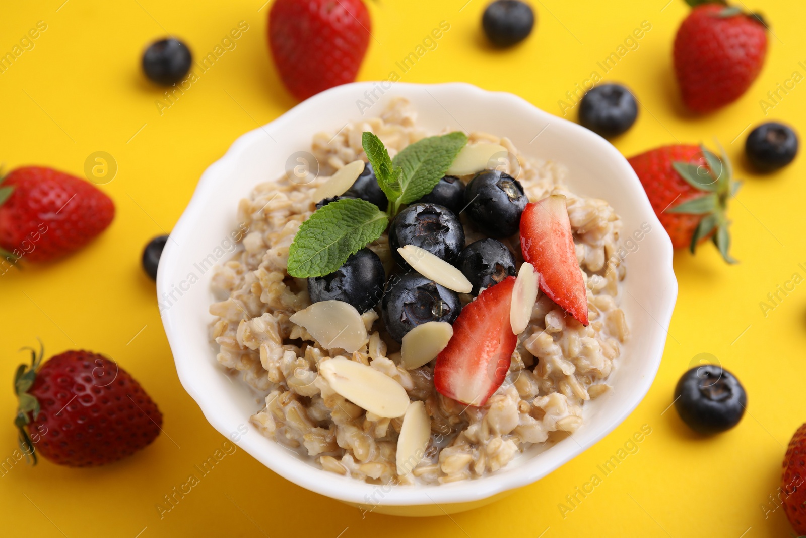 Photo of Tasty oatmeal with strawberries, blueberries and almond petals in bowl surrounded by fresh berries on yellow background