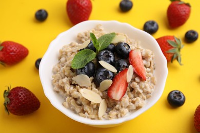 Photo of Tasty oatmeal with strawberries, blueberries and almond petals in bowl surrounded by fresh berries on yellow background