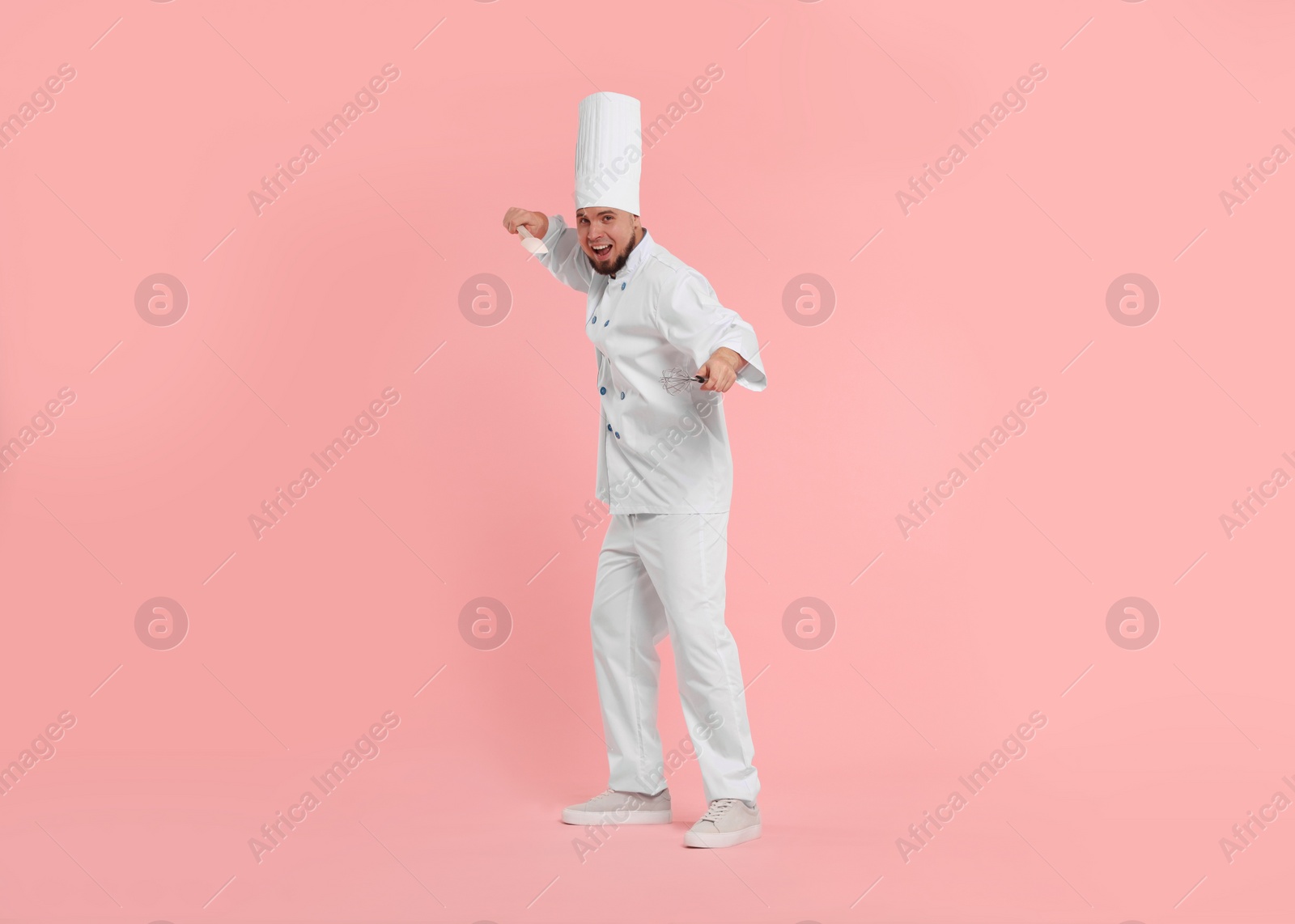 Photo of Happy professional confectioner in uniform holding spatula and whisk on pink background